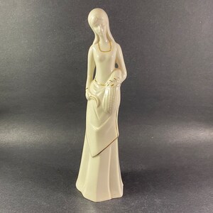 May include: A white porcelain figurine of a woman in a long dress with gold accents. She is holding a sheaf of wheat in her right hand.
