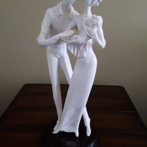 May include: White porcelain figurine of a couple holding a baby. The couple is standing on a dark brown base.