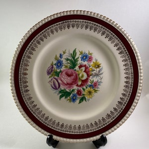 Puede incluir: Un plato de cena de cerámica blanca con un borde burdeos y dorado. El centro del plato presenta un diseño floral con flores rosas, amarillas y azules.