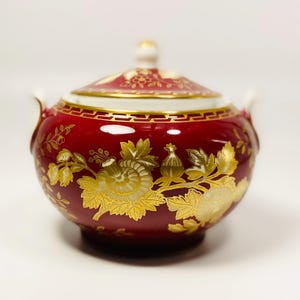 Op de afbeelding: Een rode suikerpot met een deksel, versierd met gouden bloemmotieven. De pot heeft een ronde vorm, kleine handvatten en een gouden rand. Het bloemmotief omvat bloemen en bladeren. Het deksel heeft een kleine, witte knop.