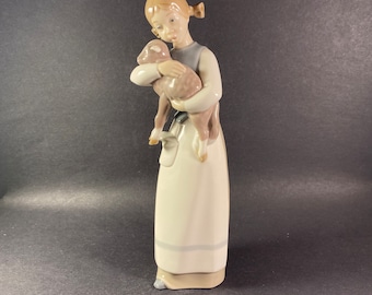 May include: A porcelain figurine of a young girl holding a lamb in her arms. The girl is wearing a white dress with a blue and grey trim. The lamb is brown and white.