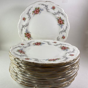 Puede incluir: Una pila de 10 platos de cena de porcelana blanca con un borde dorado y un diseño floral. Los platos presentan un delicado patrón floral gris con flores rosas y azules.