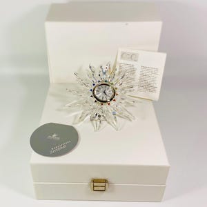 Könnte beinhalten: Eine Kristalluhr in Form einer Sonne mit weißem Zifferblatt und schwarzen Zeigern. Die Uhr befindet sich in einer weißen Box mit einem Echtheitszertifikat. Die Box hat einen goldenen Verschluss.
