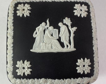 May include: Black and white ceramic trinket box with a raised design of three figures and a tree. The box has a square shape and a white border.