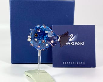 May include: A faceted blue crystal pufferfish figurine with small protruding spikes, mounted on a clear rod and a light green base. The set includes a dark blue box and a square certificate card featuring the Swarovski logo and text.