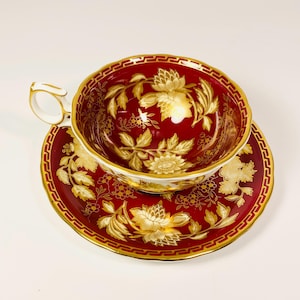 Op de afbeelding: Een rode theekop en schotel set met gouden bloemmotieven. De theekop heeft een wit handvat en een gouden rand. De schotel heeft een bijpassend ontwerp. Waarschijnlijk een vintage of antieke set.