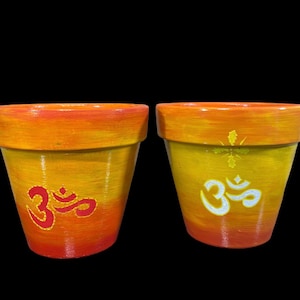 May include: Two terracotta flower pots with a gradient of orange and yellow paint. One pot has a red Om symbol painted on it, and the other has a white Om symbol painted on it.