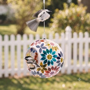 May include: A spherical bird feeder featuring a white base decorated with colorful mosaic flower patterns in shades of blue, yellow, and red. The feeder has a circular opening where a small black and white bird is perched inside.