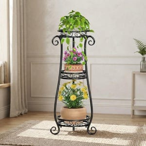 May include: A tall, black metal plant stand featuring three circular tiers with scrollwork detailing. The stand holds three potted plants: a trailing green vine on the top, pink flowers in the middle, and yellow flowers in the bottom pot.