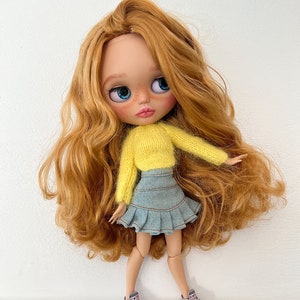 Può includere: Una bambola con lunghi capelli ondulati castano chiaro, che indossa un maglione giallo e una gonna di jeans blu. La bambola indossa scarpe da ginnastica rosa e bianche e ha gli occhi azzurri con le lentiggini.