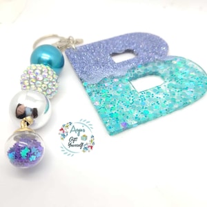 May include: A blue and purple glitter resin letter B keychain with a silver and blue beaded charm. The charm has a clear glass bead with purple glitter stars inside.