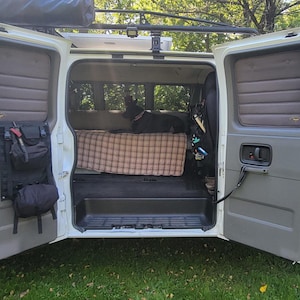 Op de afbeelding: Een witte bestelwagen met de deuren open, waardoor een bed met een geruit dekbed en een zwarte hond in het interieur zichtbaar zijn. De bestelwagen heeft een dakrek en een sticker met een bergketen aan de zijkant.