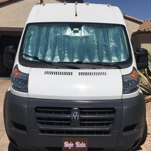 May include: White van with a silver sunshade on the windscreen. The van has a black grille with a Ram logo and a number plate that reads "Mojo Risin".