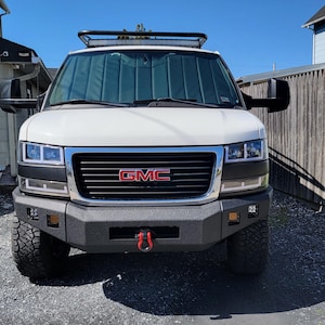 May include: White GMC van with a black front bumper, a black grille, and a red GMC logo. The van has a roof rack and a black sunshade.