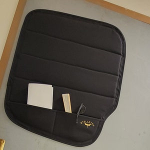 May include: Black padded organiser with a pocket for storing items such as a notebook, book, and glasses. The organiser has a gold embroidered logo that says "Stelletek" with a dragonfly design.