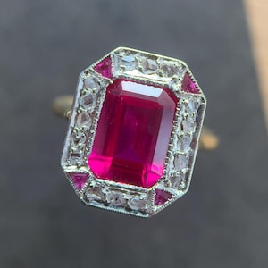 Può includere: Un anello d'oro con una grande gemma rossa rettangolare, circondata da pietre bianche e rosse più piccole. L'incastonatura dell'anello è dettagliata con un metallo color argento, creando un contrasto sorprendente con la fascia d'oro.