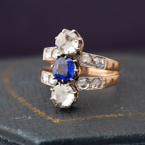 May include: A gold ring with a blue sapphire and two round diamonds. The ring is set on a dark blue velvet box.