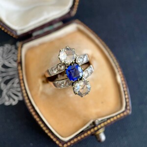 May include: A vintage ring with a blue sapphire and two white diamonds set in a silver band. The ring is displayed in a velvet-lined jewelry box.