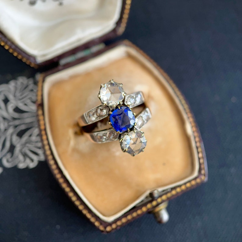 May include: A vintage ring with a blue sapphire and two white diamonds set in a silver band. The ring is displayed in a velvet-lined jewelry box.