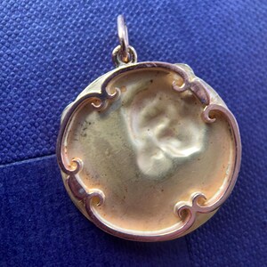 May include: A gold-colored pendant with a round, ornate frame. The frame has a decorative design with swirls and curves. The pendant is blank and ready for personalization.