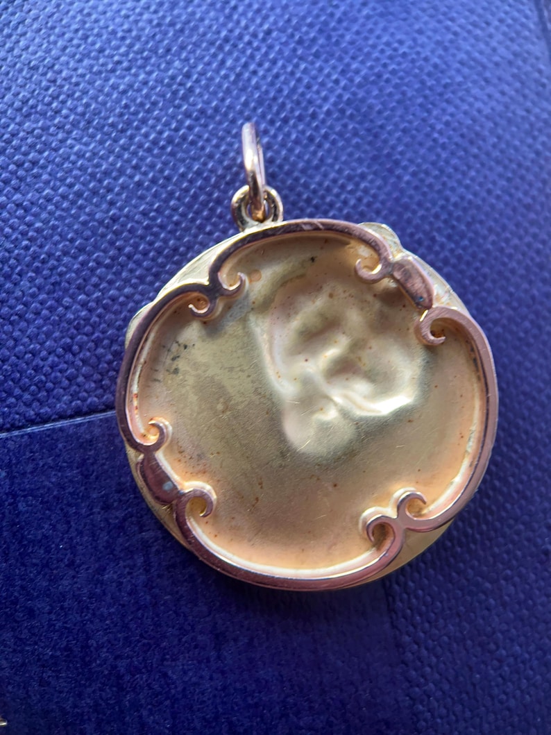 May include: A gold-colored pendant with a round, ornate frame. The frame has a decorative design with swirls and curves. The pendant is blank and ready for personalization.