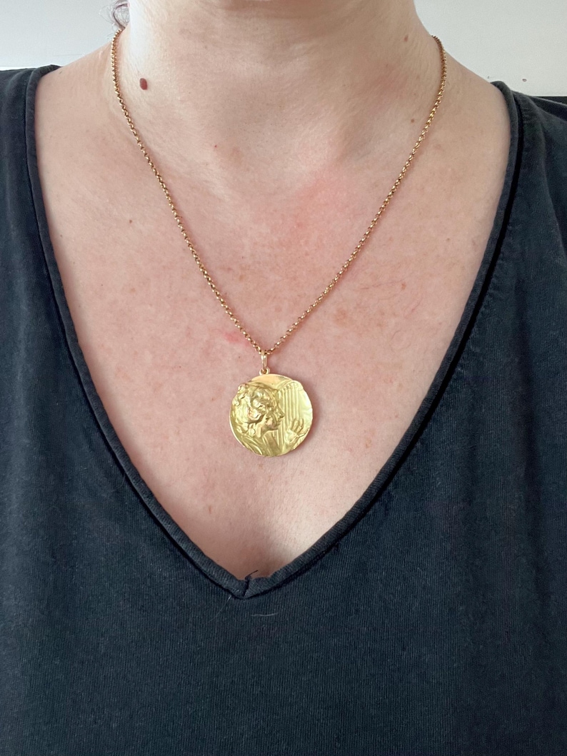 May include: A gold chain necklace with a large, round pendant featuring a detailed embossed design of a woman's face and a harp.