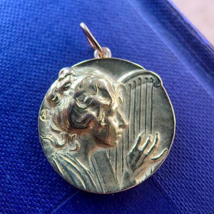 May include: A gold-toned pendant featuring a woman's profile playing a harp. The pendant is round and has a detailed design.