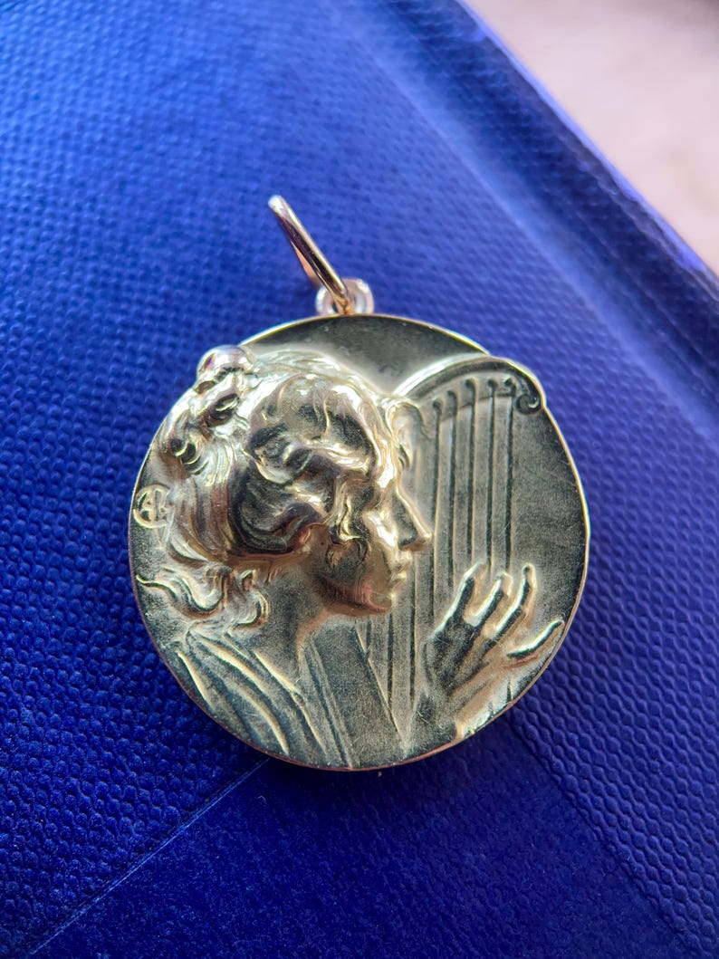 May include: A gold-toned pendant featuring a woman's profile playing a harp. The pendant is round and has a detailed design.