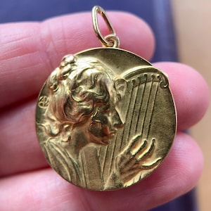 May include: A gold-toned pendant featuring a detailed relief of a woman playing a harp. The pendant is oval-shaped and has a loop at the top for attaching to a chain.