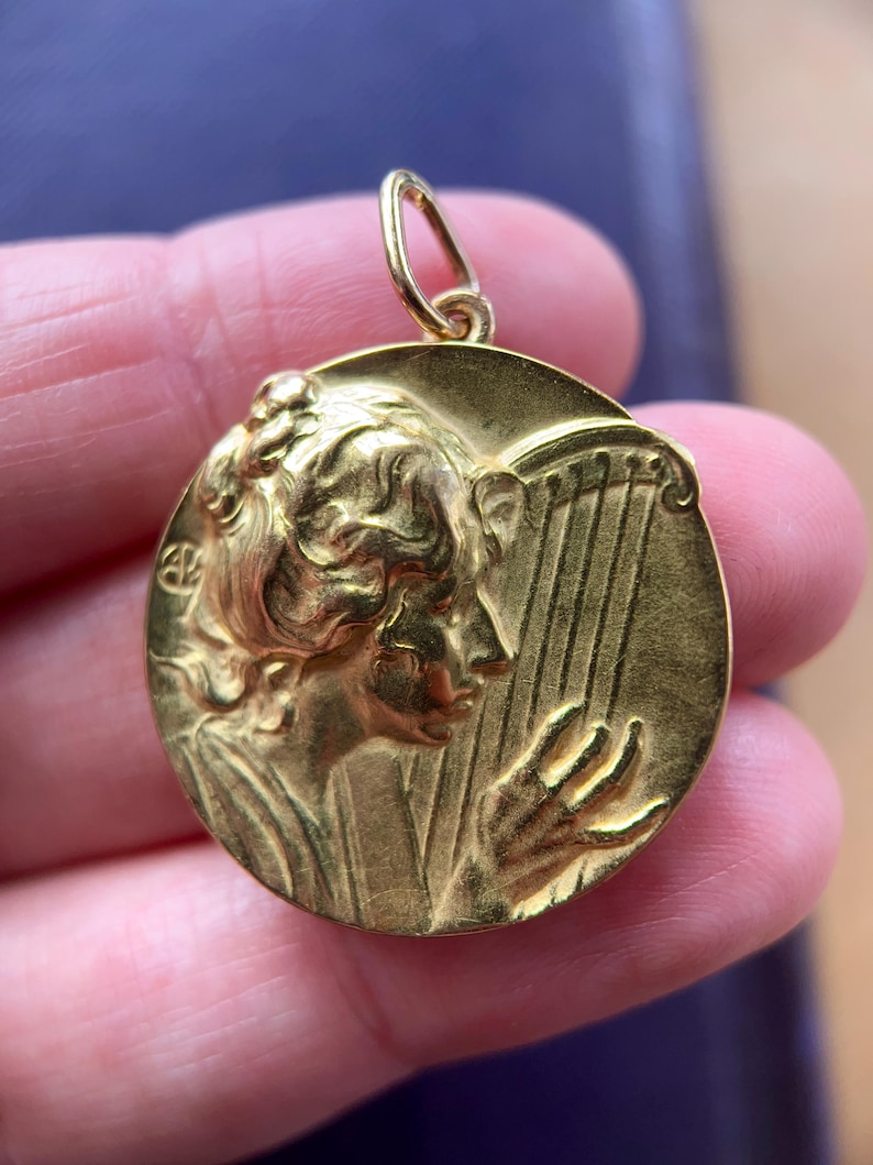 May include: A gold-toned pendant featuring a detailed relief of a woman playing a harp. The pendant is oval-shaped and has a loop at the top for attaching to a chain.