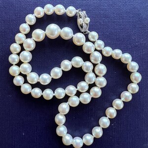 May include: A strand of white pearls with a silver clasp. The pearls are strung on a white thread and are arranged in a circular pattern. The clasp is a small, silver piece with a diamond-shaped design.