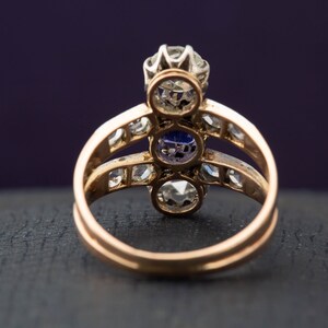 May include: A gold ring with three round gemstones. The center stone is a large, clear diamond. The two stones on either side are smaller and have a purple hue. The ring is set with smaller diamonds around the stones.
