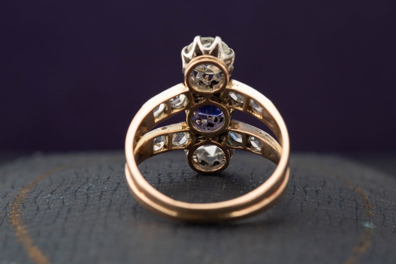 May include: A gold ring with three round gemstones. The center stone is a large, clear diamond. The two stones on either side are smaller and have a purple hue. The ring is set with smaller diamonds around the stones.