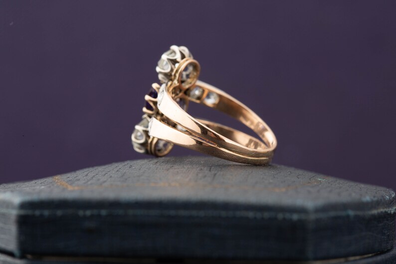 May include: A gold ring with a cluster of purple and white gemstones. The ring is set on a black velvet box.