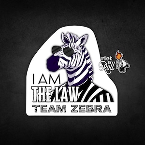May include: A black and white zebra wearing sunglasses and a black and white striped shirt. The zebra has the text "I AM THE LAW TEAM ZEBRA" printed below it. The image also includes a logo for "Riot & Roll" in the upper right corner.