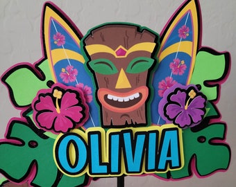 May include: A colorful cake topper featuring a tiki mask, surfboards, tropical flowers, and the name "Olivia".
