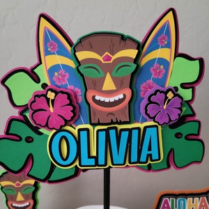 May include: A colorful cake topper featuring a tiki mask, surfboards, tropical flowers, and the name "Olivia".