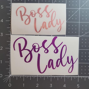 May include: Two vinyl decals with the text "Boss Lady" in a cursive font. The top decal is rose gold and the bottom decal is purple.
