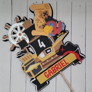 May include: A yellow, brown, and black pirate ship cake topper with a red parrot, a treasure chest, a steering wheel, and a black pirate hat. The topper also includes the number 4 and the name Gabriel.