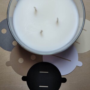 Overhead view of candle dust covers in black, gray, white, and cream on a wood background. Standard and wood wick options.