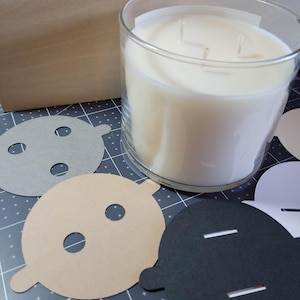 Overview of candle dust covers in a variety of color options, including black, white, cream, gray, and kraft brown. A silver tin candle is featured with a black dust cover in standard wick as an example.