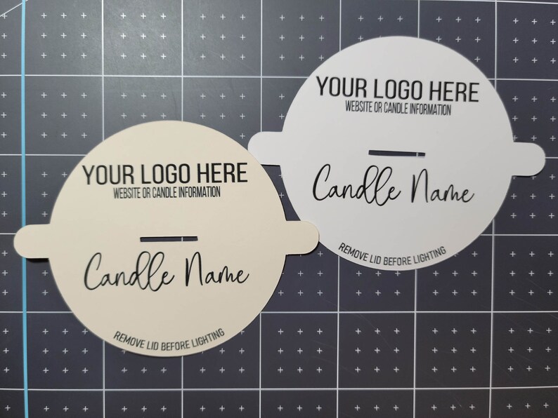 Another photo of two custom candle dust cover examples - one in cream, one in white, both with wood wicks and basic sample text on top of a measuring grid for size.