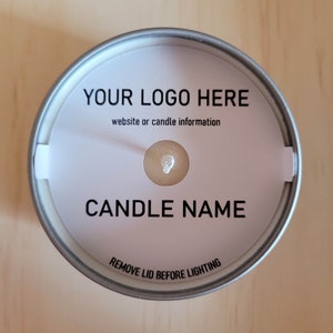 May include: A silver candle tin with a white label. The label says "YOUR LOGO HERE" and "website or candle information". Below that, it says "CANDLE NAME" and "REMOVE LID BEFORE LIGHTING".