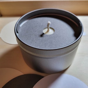 Overview of candle dust covers in a variety of color options, including black, white, cream, gray, and kraft brown. Focus is on a silver tin candle with a gray dust cover and a standard wick.