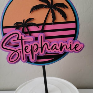 Retro style sunset cake topper in orange, hot pink, purple, neon blue, and black on a white cake with a black cake stick.