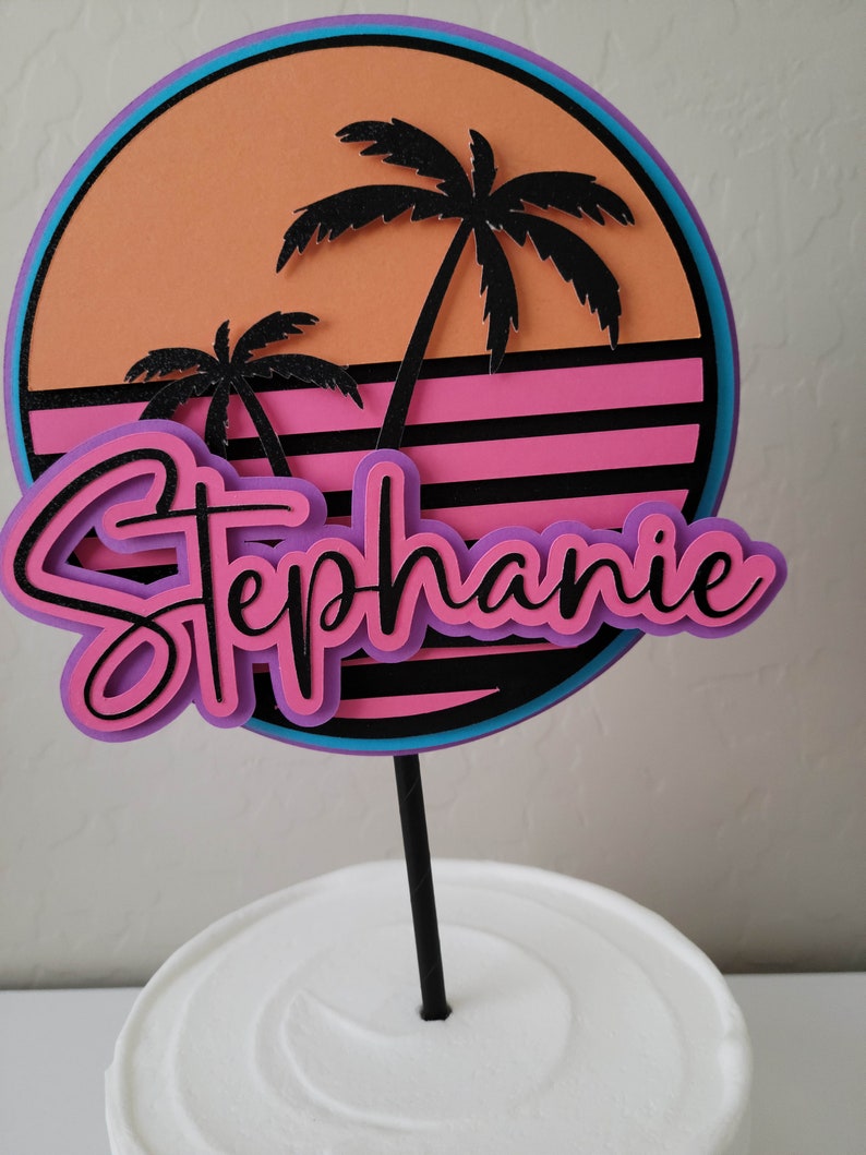 Retro style sunset cake topper in orange, hot pink, purple, neon blue, and black on a white cake with a black cake stick.