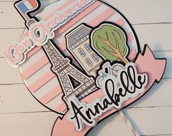 May include: A pink and white striped cake topper with a black silhouette of the Eiffel Tower, a small building, a tree, and two bicycles. The topper also includes the text "Bon Anniversaire" and "Annabelle".