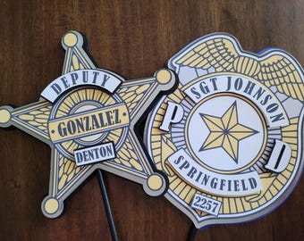 May include: Two law enforcement badges, a star-shaped Deputy badge and a shield-shaped Police badge. The Deputy badge has the name "Gonzalez" and "Denton". The Police badge has the name "Sgt Johnson" and "Springfield". Both badges are yellow, white, and gray.