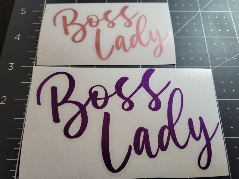 Photo featuring two examples of the Boss Lady vinyl decal - rose gold in 4-inches and purple sparkle in 5-inches - on a gray measuring mat for size.