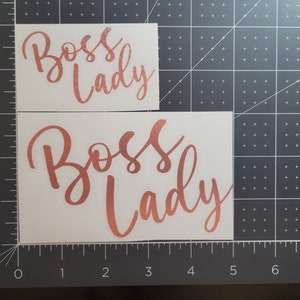 Photo featuring two examples of the Boss Lady vinyl decal - rose gold in 4-inches and 5-inches - on a gray measuring mat for size.
